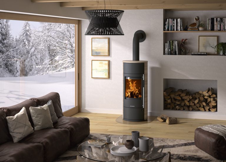 ORANIER Kaminofen Polar Neo W+ Stahl Schwarz Grappa in einem Wohnzimmer mit bequemer Couch. Holzlegefach links im Bild. Schneelandschaft durch Große Fenster zu sehen.