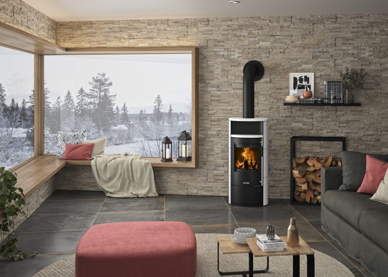 Bequemes Wohnzimmer mit Fensterbank zum Sitzen. Kaminofen mit modernem Metall Holzlegefach. Schneelandschaft durch Fenster zu sehen.