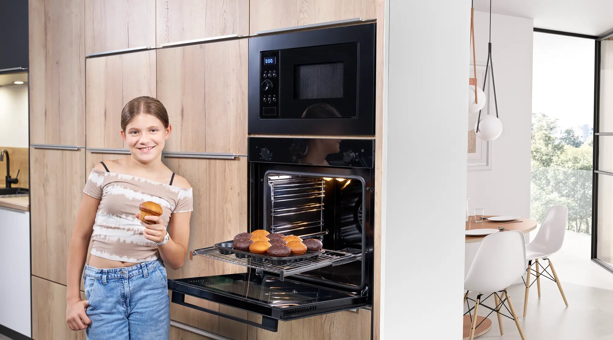 Ein Mädchen steht vor einem ORANIER EBS220-Einbaubackofen und holt einen Muffin aus dem Backofen und über dem Backofen ist eine ORANIER HMG360-Mikrowelle.