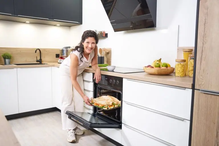 Eine junge Frau in einer modernen hellen Küche, die ein Backblech mit Gemüse aus einem ORANIER EBH380- Herdset