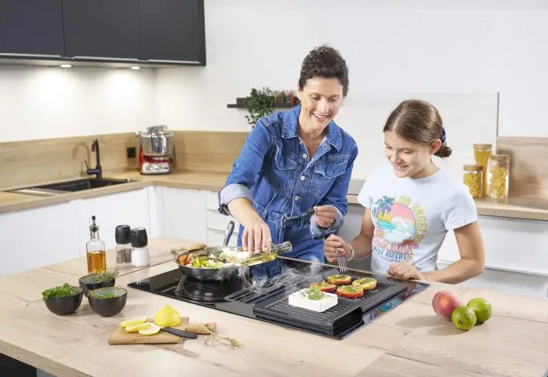 Eine junge Frau und ein Mädchen, die in einer modernen Küche auf der Kochinsel kochen, mit einem ORANIER KFA590-Kochfeldabzug und mit einer Grillplatte von ORANIER KGP260.