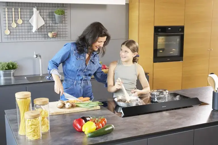 Eine junge Frau und ein Mädchen, die in einer modernen Küche auf der Kochinsel kochen, mit einem ORANIER KFL2094-Kochfeldabzug und im Hintergrund ein ORANIER EBS420-Einbaubackofen