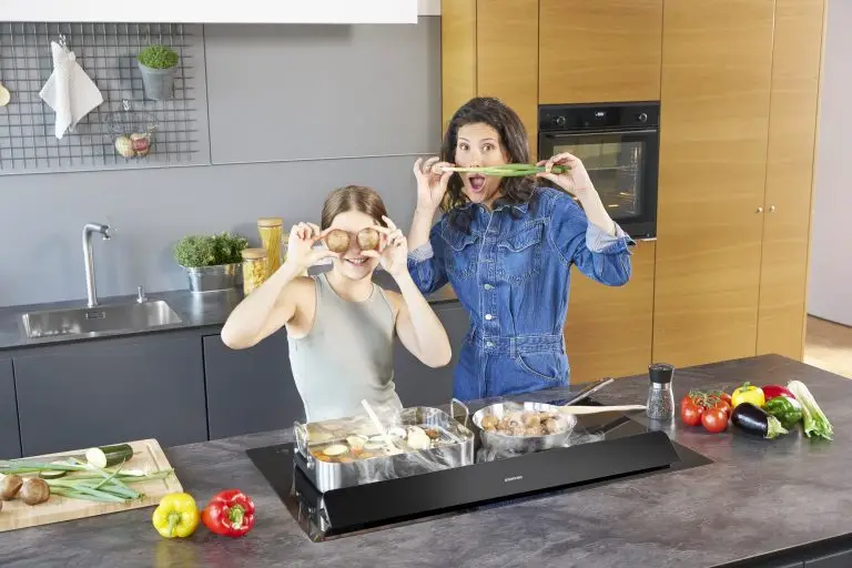 Eine junge Frau und ein Mädchen, die in einer modernen Küche auf der Kochinsel kochen, mit einem ORANIER KFA590-Kochfeldabzug und im Hintergrund ein ORANIER EBS420-Einbaubackofen