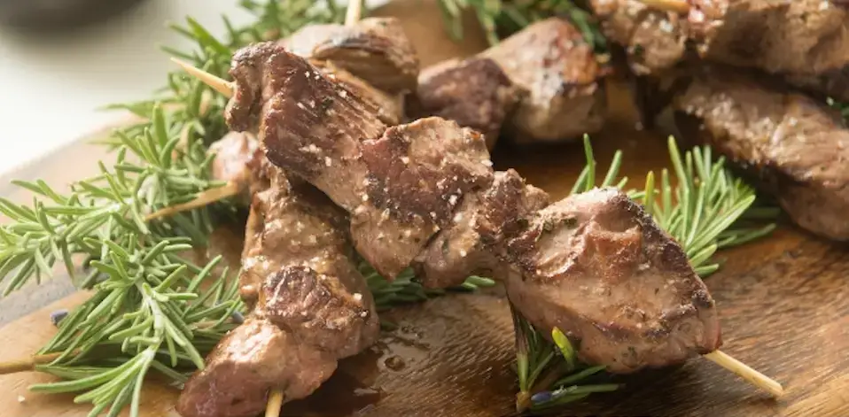 Auf dem Bild sieht man Honig-Kräuter-Lamm-Spieße auf einem Holzbrett angerichtet